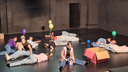 Jugendliche liegen auf der Bühne und halten Luftballons. In der Mitte sitzt ein Mädchen vor einem Springteufel.